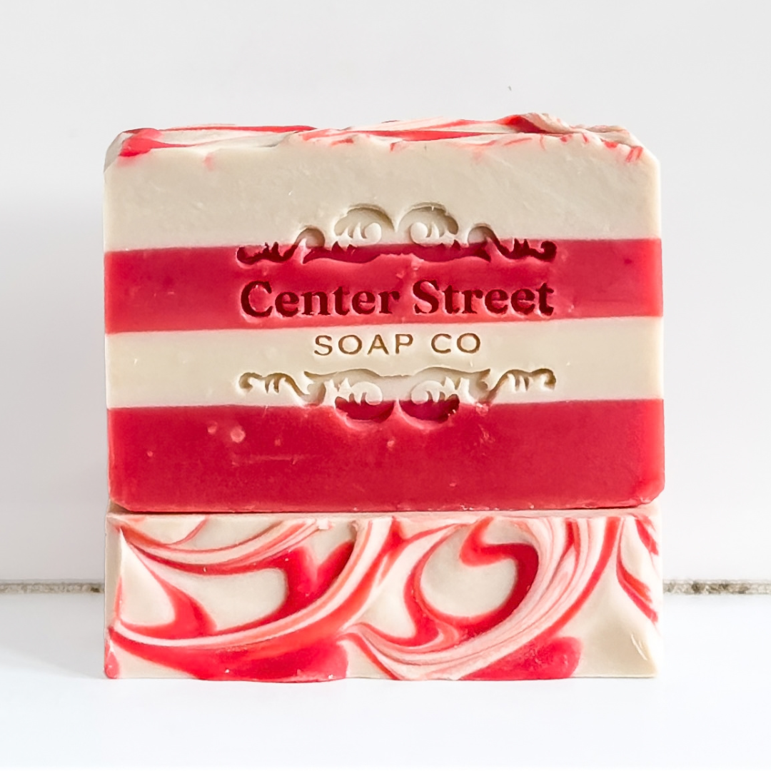 A bar of handmade soap with red and cream horizontal stripes and a swirled pattern on the bottom half. The brand name 'Center Street SOAP CO' is embossed on the front, set against a plain white background.