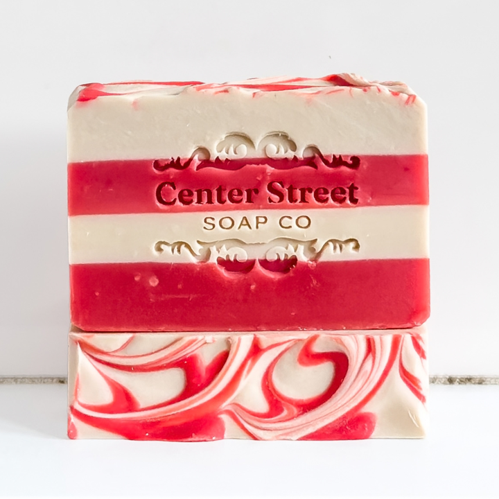 A bar of handmade soap with red and cream horizontal stripes and a swirled pattern on the bottom half. The brand name &