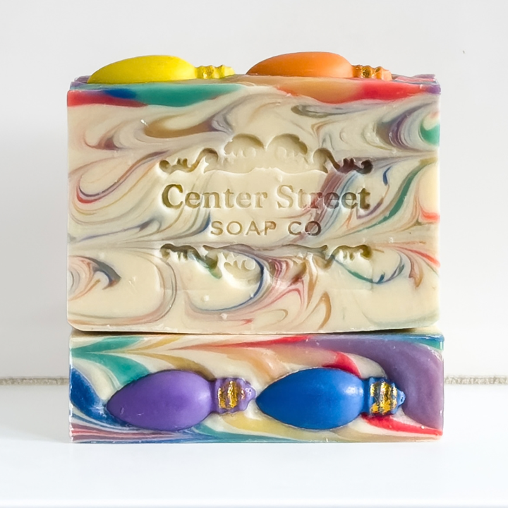 Two rectangular bars of marbled handmade soap with colorful swirls and decorative embossed lettering, shown on a white background. The top bar is cream with multicolor swirls and yellow/orange tops; the bottom bar has purple, blue, and red swirls with gold-toned bead details.