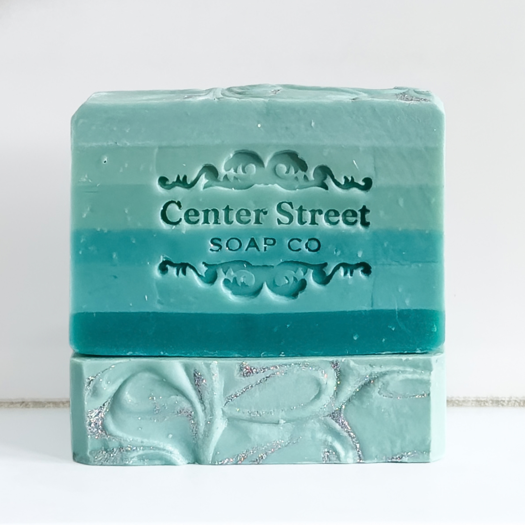 A handmade bar of soap in a mint green and teal gradient with silver glitter swirls, embossed with the brand name 'Center Street SOAP CO'.