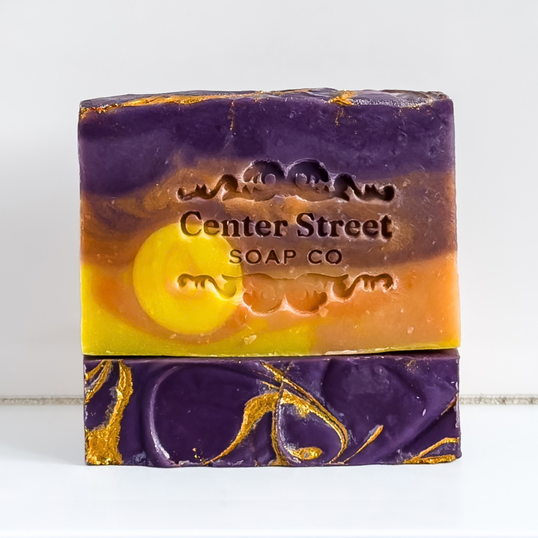 A rectangular bar of handmade soap with a marbled design in purple, orange, yellow, and gold foil, embossed with the brand name 'Center Street SOAP CO'.