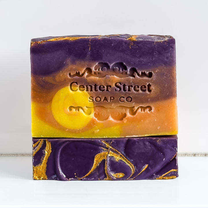 A rectangular bar of handmade soap with a marbled design in purple, orange, yellow, and gold foil, embossed with the brand name &