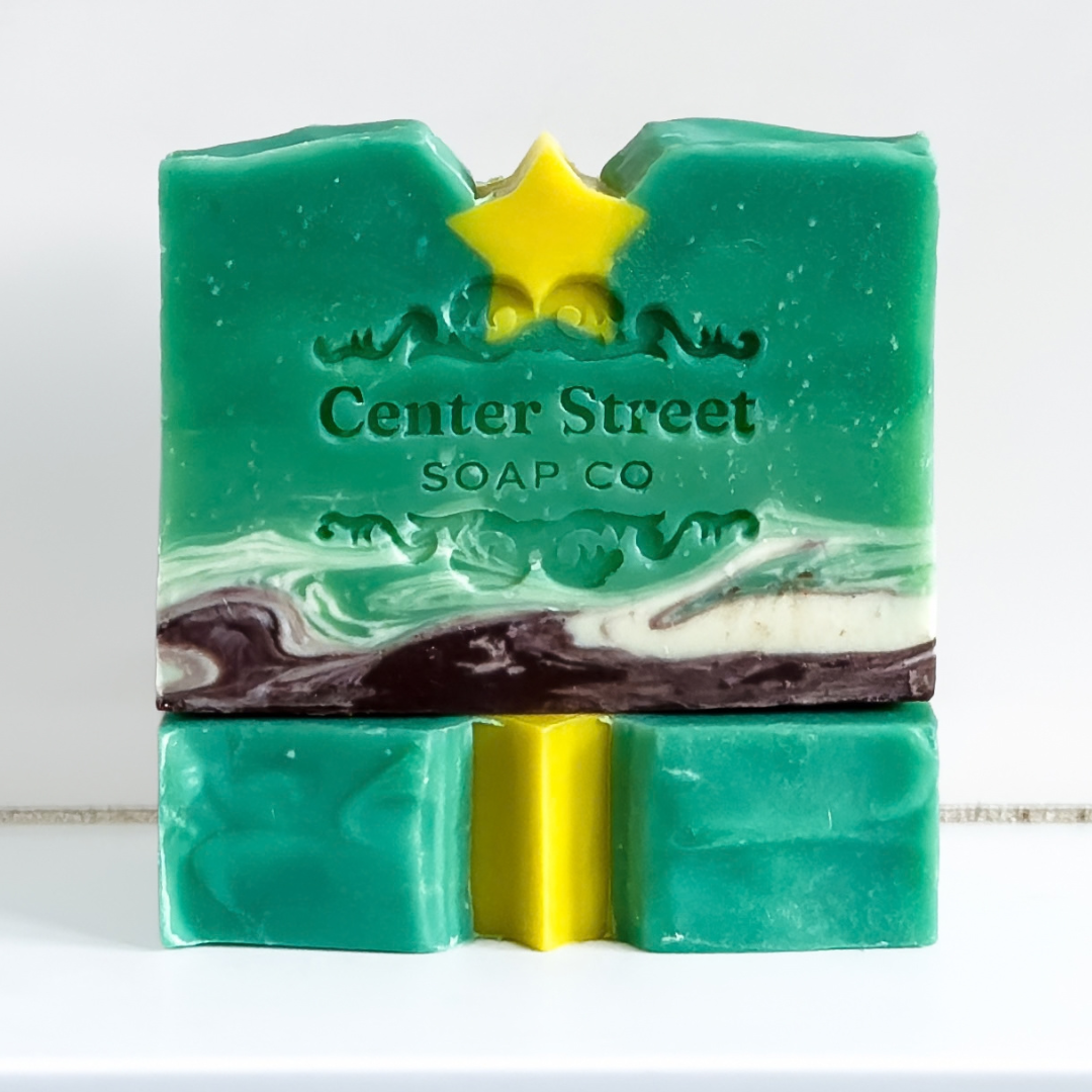 A handmade bar of soap from Center Street Soap Co, featuring a marbled green, white, and dark brown pattern with a bright yellow star detail on top, displayed on a white surface against a white background.